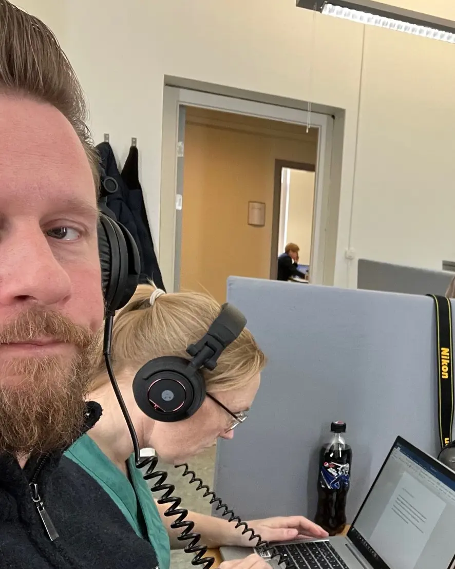 Portrett av en mann med hodetelefoner. I bakgrunnen ser du en kvinne som bøyer seg fram foran en PC
