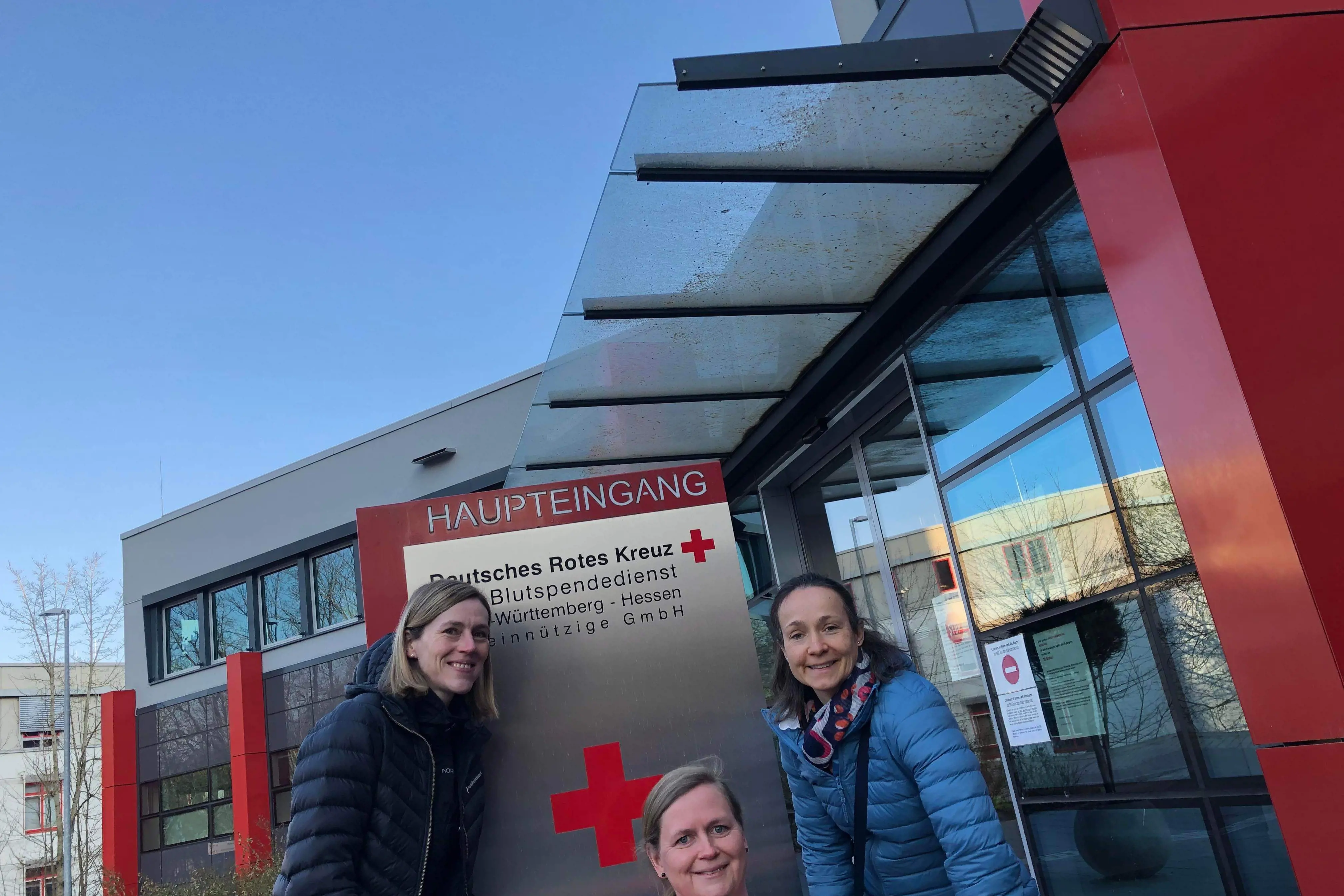 Three persons in front of Red Cross sign. Photo