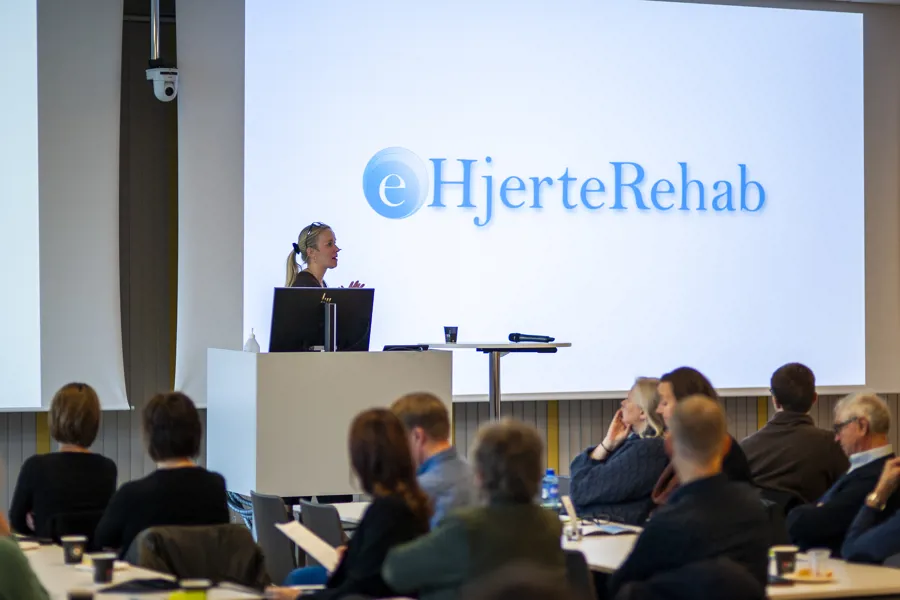 Prosjektleiar Tone Norekvål på scenen foran forsamling i konferanselokale, presentasjon om ehjerterehabilitering på skjerm. Foto