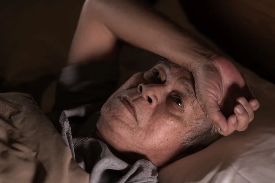 Eldre person ligger i seng, tankefullt blikk. Foto