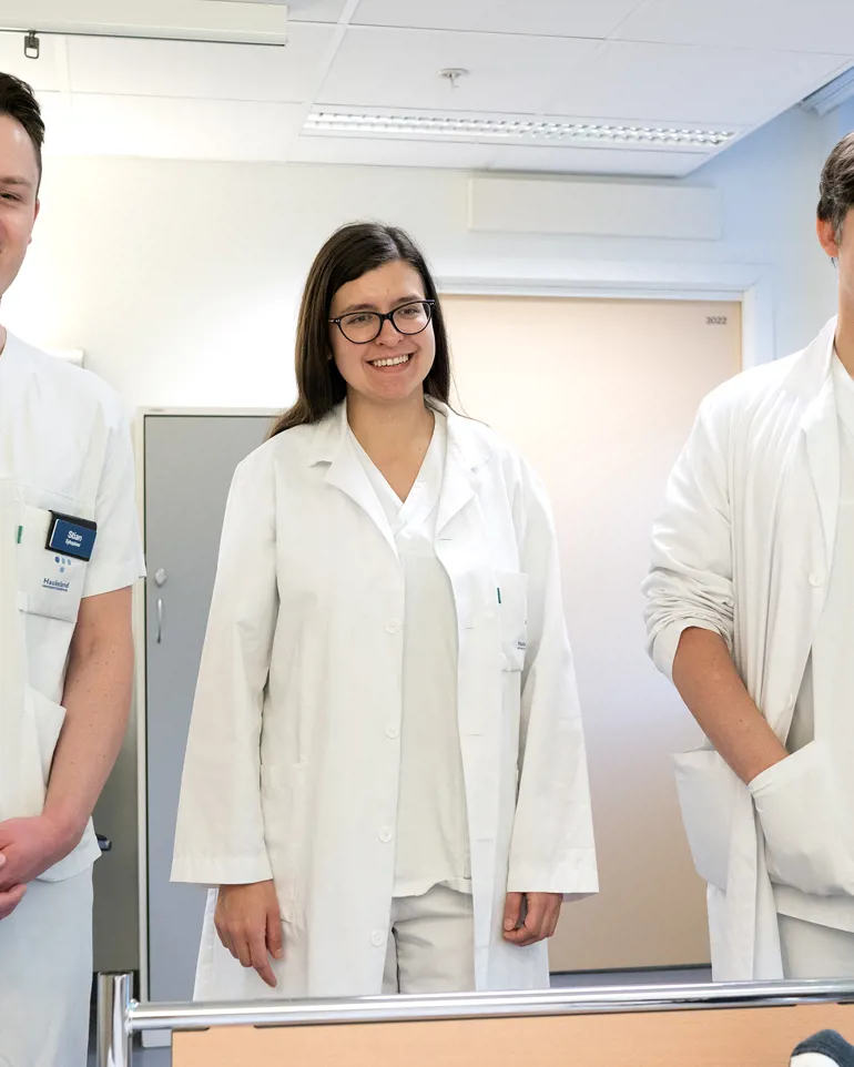 Gruppebilde to smilende menn og en kvinne, kledd i hvite uniformer i sykehus. Foto
