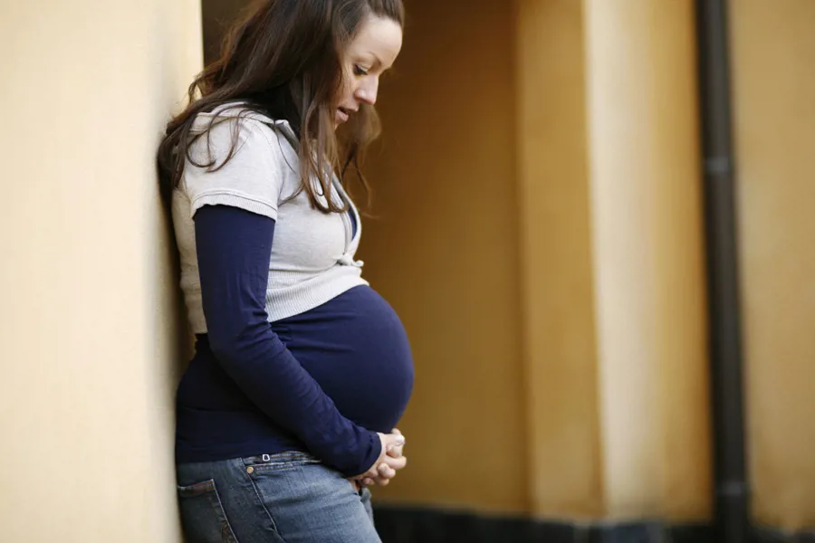 Gravid kvinne lent inntil vegg, ser ned på magen. Foto