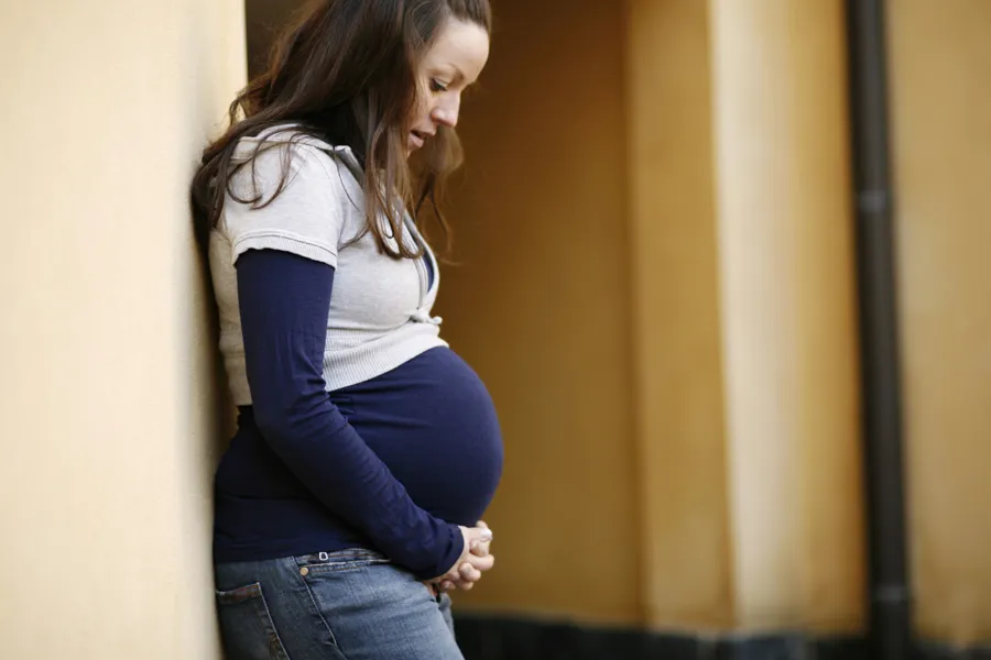 Gravid kvinne lent inntil vegg, ser ned på magen. Foto