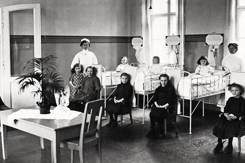 Interiør sykestue med barnepasienter i senger og sittende på stoler, to sykepleiere. Foto