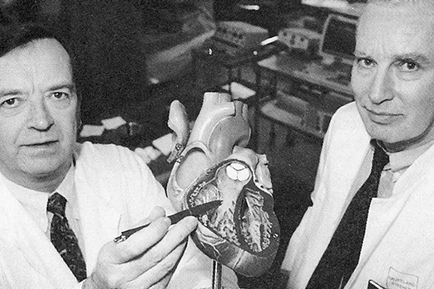 To menn i hvite frakker med modell av hjertet, ene peker med kulepenn på hjertemodellen, i undersøkelsesrom. Foto
