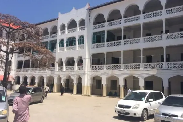 Eksteriør sykehusbygg Mnazi Mmoja Hospital i Zanzibar, parkeringsplass med tre, biler og folk. Foto