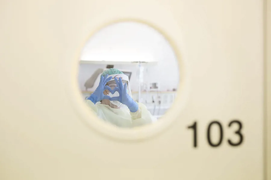 Foto: Vindu på dør hvor sykepleier i smittevernutstyr former fingrene som ett hjerte
