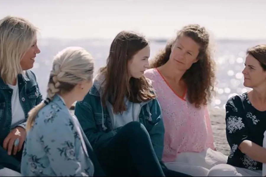 Ung jente sitter omringet av fire kvinner på et svaberg med havet i bakgrunnen. Foto
