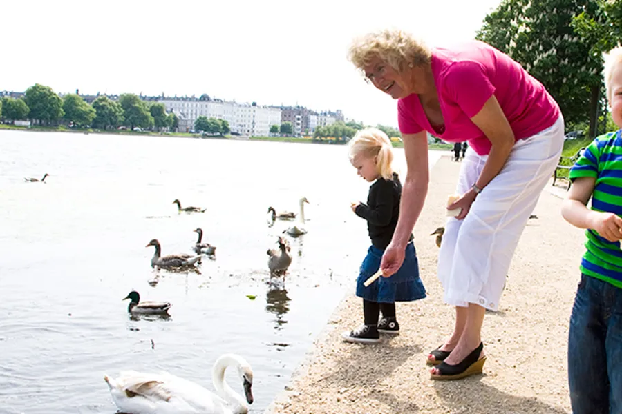 Eldre kvinne mater endre ved vann sammen med liten gutt og jente. Foto