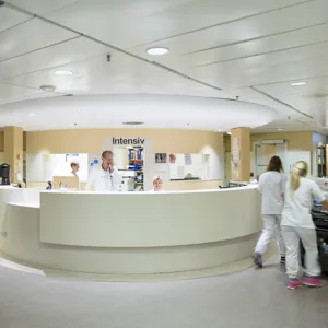 Viser en intensivsentral hvor en kvinne sitter å smiler bak skranken, mens en mann står og snakker i telefonen.