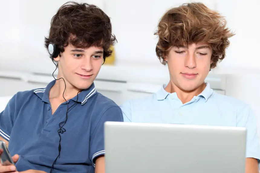 To unge gutter sitter i sofa, en med mobil og hodetelefoner og den andre med laptop i fanget. Foto