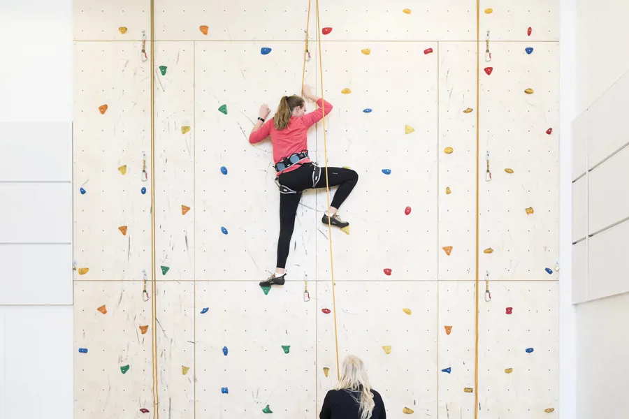 Kvinne i treningstøy og sikkerhetsutstyr klatrer i klatrevegg mens kvinne passer på. Foto
