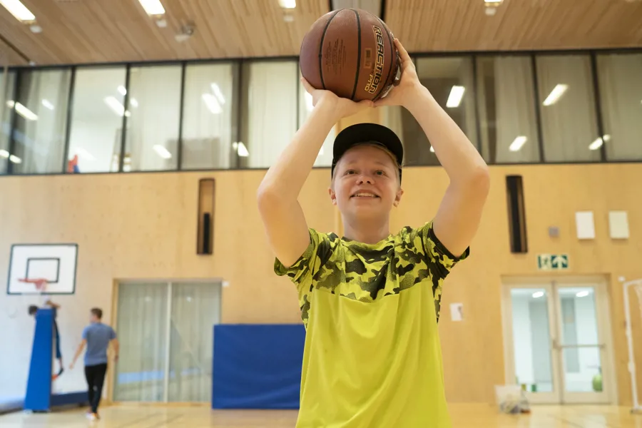 Ung gutt med gul tskjorte og caps med basketball i gymsal. Foto