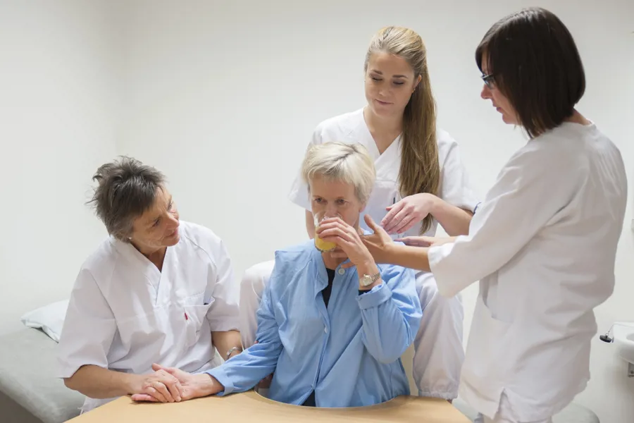 Tre damer i hvitt sykehustøy hjelper sittende pasient med å drikke