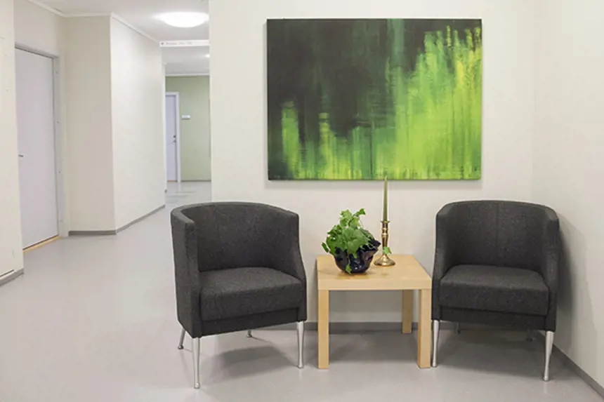 Interiør lite venterom med grønt bilde på veggen, to stoler, bord med lysestake og plante. Foto
