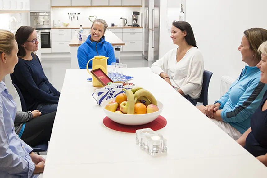 Gruppesamtale. Seks kvinner sitter rundt møtebord med kaffe og frukt. Foto.
