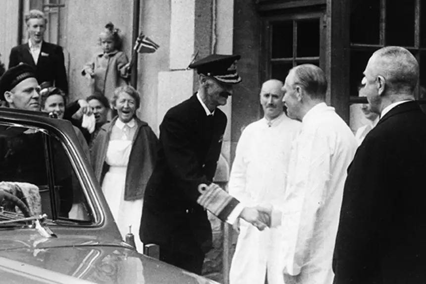 Kong Håkon hilser på leger, sykepleiere, barn med flagg utenfor inngang. Foto